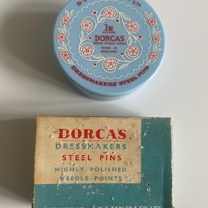 Könnte beinhalten: Zwei Vintage-Behälter mit Dorcas-Schneiderstahlnadeln. Einer ist eine hellblaue Dose mit floralen Designs und dem Text "DORCAS" und "DRESSMAKERS' STEEL PINS". Der andere ist eine rechteckige Schachtel mit ähnlichem Text.
