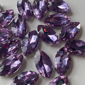 May include: A collection of marquise-shaped, faceted, purple rhinestones. Each stone has a silver-colored metal setting. The stones are arranged on a white surface, reflecting light and creating a sparkling effect. These rhinestones are suitable for various craft projects.