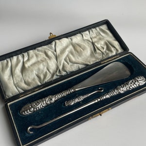 Könnte beinhalten: Ein antikes Set aus silbernem Schuhanzieher, Knopfloch und Schuhanzieher in einem schwarzen Etui mit Samtfutter. Die Werkzeuge haben verzierte, detaillierte Griffe. Das Etui ist geöffnet und zeigt die Werkzeuge vor einem hellgrauen Hintergrund.