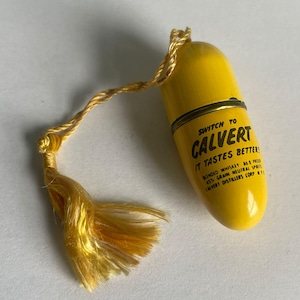 May include: A yellow pill-shaped container with a gold tassel. The container has the text "SWITCH TO CALVERT IT TASTES BETTER!" printed on it. The tassel is made of gold-colored threads.