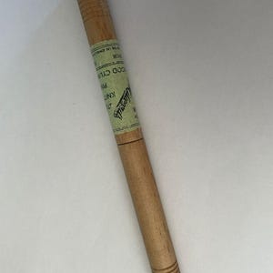 以下が含まれることがあります： 「Wood Cylinder Pin」と書かれたラベルが付いた木製の編み針。 針は薄茶色で、両端に溝のディテールがあります。 ラベルは緑と白で、テキストが部分的に表示されています。