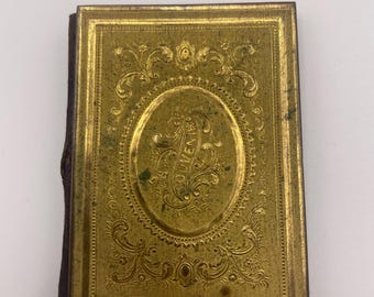May include: A small, gold-coloured book with an ornate, embossed design on the cover. The design features a central oval with the word "OLEARY" inscribed inside. The book is bound in brown leather.