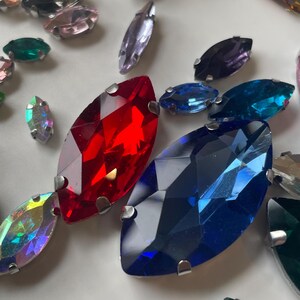 May include: Assortment of colorful, marquise-shaped rhinestones in various colors, including red, blue, green, and iridescent. Each rhinestone has a silver metal backing. The rhinestones are scattered on a white surface.
