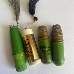 May include: A selection of vintage thimble holders and thimbles. The thimble holders are green and gold, with tassels. One holder features a crest design. Two thimbles are gold-toned, and one has text that reads "PHARMACIE PRINCIPAL TOURS".