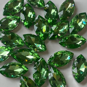 May include: A collection of marquise-shaped, faceted green gemstones. Each gem has a pointed oval shape and is set with small silver-colored prongs. The gems are a vibrant, light green color and reflect light.