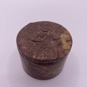 May include: A small, round, carved stone box with a lid featuring a detailed angel design. The box is made of brown and white marbled stone.