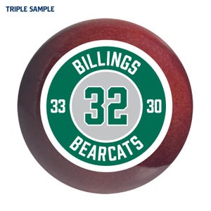 May include: A red and white baseball with a green circle around it. The circle says "Billings Bearcats" with the number 32 in the center. The numbers 33 and 30 are on either side of the number 32.