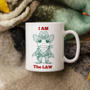 May include: White ceramic mug with a cartoon frog wearing a sheriff's hat and a red bandana. The mug has the text "I AM THE LAW" printed on it.