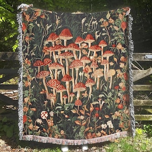 May include: A tapestry throw blanket with a dark green background and a pattern of red and white mushrooms. The mushrooms are arranged in a cluster and are surrounded by green leaves and flowers.