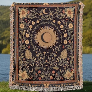 May include: A woven tapestry throw blanket with a dark blue background and a floral pattern. The center of the blanket features a sun and moon design.