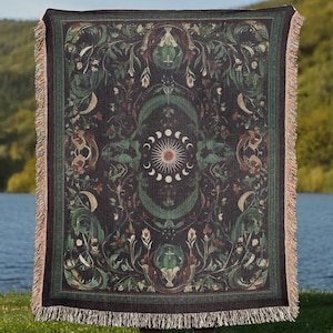 Peut inclure: Une couverture en tapisserie avec un motif de soleil central, de lune et d'étoiles, entouré de motifs floraux. Les couleurs dominantes sont le vert foncé, le noir et le marron.