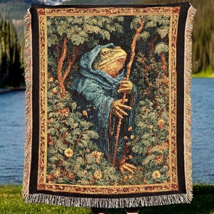 May include: Woven tapestry blanket featuring a frog-like figure in a blue robe holding a staff, set in a forest scene. The blanket has a black border with fringe and a detailed, colorful design. The background shows a lake and mountains.