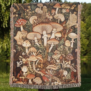 Peut inclure: Une couverture en tapisserie représentant une illustration colorée et détaillée de différents champignons dans un décor de forêt. La couverture a une bordure frangée et est faite de tissu tissé.