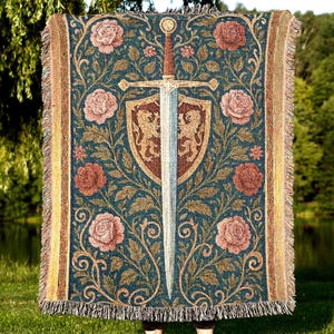 Può includere: Una coperta arazzo con un design medievale. La coperta presenta una grande spada, uno scudo con emblemi di leone e motivi floreali nei toni del blu, marrone e rosa. I bordi hanno un bordo con frange.