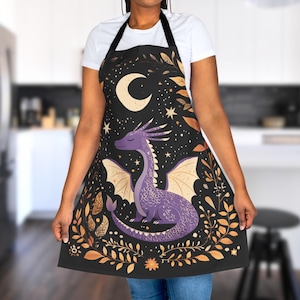 Könnte beinhalten: Eine schwarze Küchenschürze mit einem Design aus einem violetten Drachen, Mond und Sternen. Der Drache ist von goldenen floralen Akzenten umgeben.