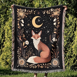 May include: A black tapestry with a gold and orange fox design. The fox is sitting under a crescent moon with stars in the background. The tapestry has a floral border with gold accents.