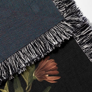 May include: Close-up of a woven throw blanket with a black and white fringe. The blanket has a dark blue background with a subtle pattern and a floral design on the reverse side.