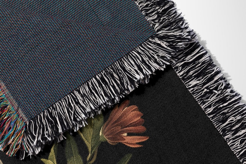 May include: Close-up of a woven throw blanket with a black and white fringe. The blanket has a dark blue background with a subtle pattern and a floral design on the reverse side.