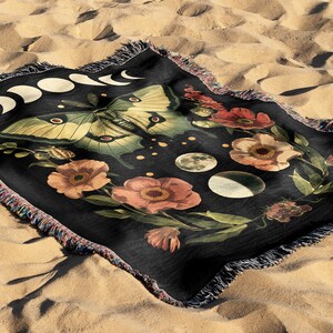 May include: A black woven tapestry throw blanket featuring a moon phases design, a green and yellow butterfly, and pink and red flowers. The background is black with white dots.