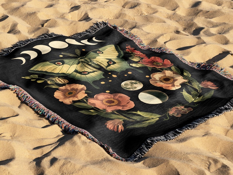 May include: A black woven tapestry throw blanket featuring a moon phases design, a green and yellow butterfly, and pink and red flowers. The background is black with white dots.