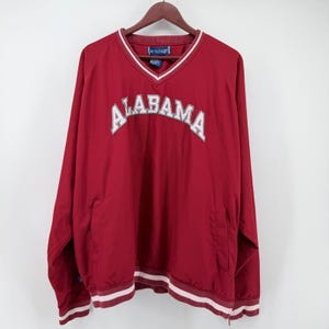 May include: A deep red long-sleeve pullover windbreaker featuring a V-neckline with white and red striped trim. The word ALABAMA is embroidered in white with a grey outline across the chest. The garment includes a blue interior brand label.