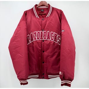 May include: A maroon satin varsity jacket featuring the word RAZORBACKS in white and maroon lettering across the chest. The jacket has a snap-button front closure, striped ribbed cuffs and collar, and a small logo on the left sleeve.