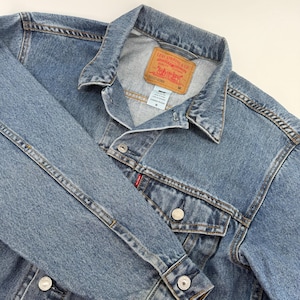 May include: A medium-wash denim jacket with a classic collar and button-up front. The jacket features silver buttons, a red Levi's tag, and a small red tag on the pocket. The jacket is a size medium.