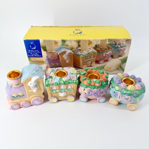 May include: A set of pastel-colored ceramic train candle holders. Each train car is decorated with floral and Easter-themed designs, including bunnies and eggs. The box reads "Rabbit & Co. Train Set Candle Holders."