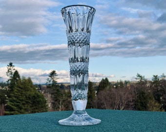 May include: A clear glass bud vase with a diamond pattern. The vase has a flared top and a wide base. It is sitting on a green surface.
