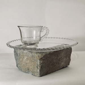 May include: A clear glass cup and saucer set with a beaded edge sits on a gray concrete block.