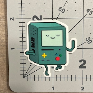 May include: A pixelated green and blue cartoon robot with a smiling face and a red button on its chest. The robot has a small screen with a white background and a black border. The robot is standing on a white background.