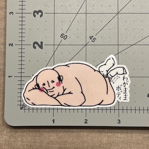 May include: A sticker featuring a cartoon illustration of a nude figure lying down, with rosy cheeks and a relaxed expression. The figure is light pink with black outlines. Japanese text is visible on the figure's sock.