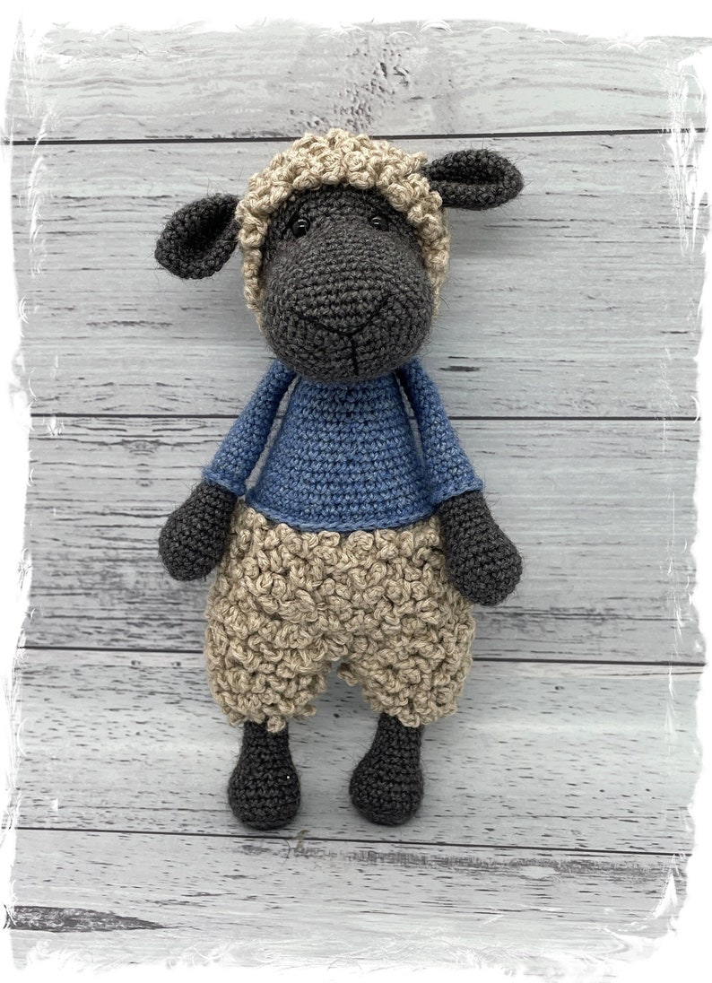 May include: A crocheted sheep toy with a black face, brown eyes, and a blue shirt. The sheep is standing on a white wooden background.