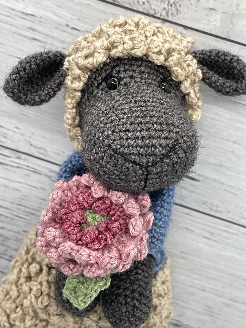 May include: A crocheted gray sheep holding a pink flower with green leaves. The sheep has a white crocheted wool hat.