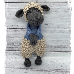 May include: A crocheted sheep toy with a black body, beige wool, and a blue sweater. The sheep is standing on a white wooden background.