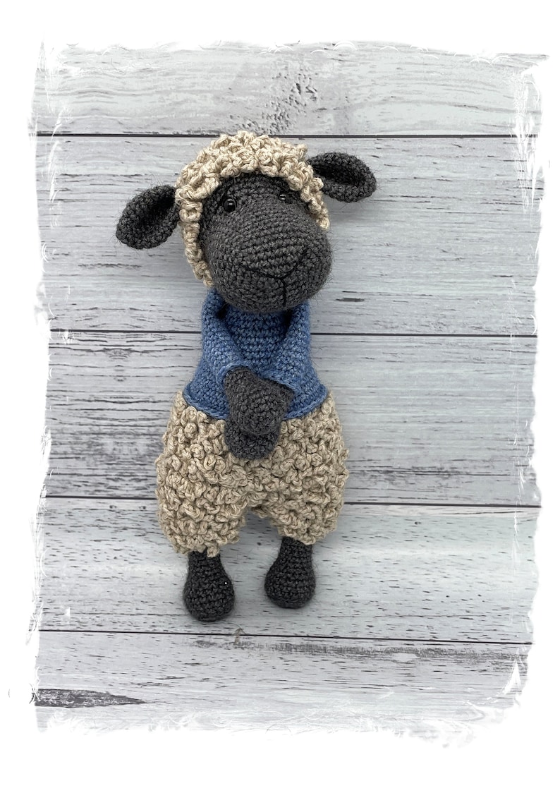 May include: A crocheted sheep toy with a black body, beige wool, and a blue sweater. The sheep is standing on a white wooden background.