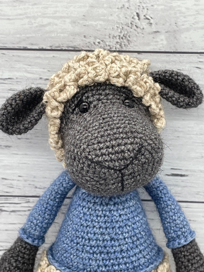 May include: A crocheted sheep toy with a gray body, black ears, and a cream-colored fleece. The sheep is wearing a blue knitted sweater.