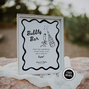 Może przedstawiać: Oprawiony znak z białym tłem i czarną obwódką, z napisem "Bubbly Bar" i instrukcjami dotyczącymi koktajlu szampańskiego. Zawiera ręcznie rysowaną ilustrację butelki i kieliszka do szampana. Znak znajduje się na kamiennej powierzchni.
