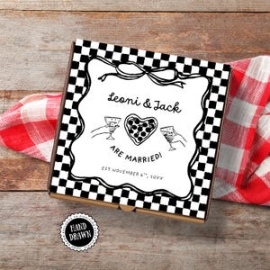 Op de afbeelding: Een vierkante pizzadoos met een zwart-wit geruit patroon. De doos heeft een handgetekend ontwerp met de tekst "Leoni & Jack ARE MARRIED!" en een hartvormige pizza. Een rood-wit geruite doek staat op de achtergrond.