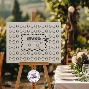 May include: A white guestbook sign on a wooden easel, with a black frame and bow, displaying the text "Louis & Samantha's GUESTBOOK." The sign has spaces for guests to draw and sign. A "Hand Drawn" sticker is also visible.