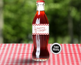 May include: A glass bottle filled with a red liquid, featuring a red cap and a label with the text "Jasmine's Ready to POP!". The label has a red and white checkered pattern. The bottle is on a red and white checkered surface.