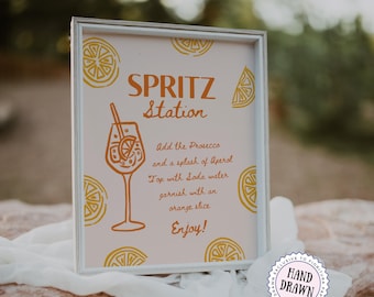 May include: A framed sign with the words "SPRITZ Station" in orange lettering. The sign includes instructions for making a spritz cocktail, with a drawing of a cocktail glass and orange slice. The sign is on a light pink background with orange citrus slice illustrations.