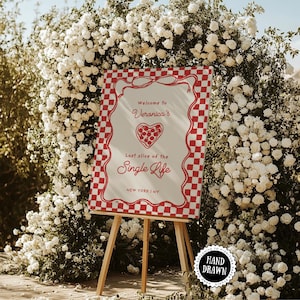 Op de afbeelding: Een houten ezel toont een bord met een rood-witte geblokte rand. Op het bord staat "Welcome to Veronica's, Last slice of the Single Life, New York, NY." Witte bloemen vormen de achtergrond.