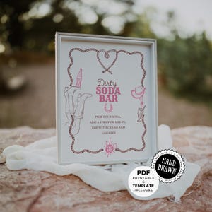 May include: A framed sign with a pink and white design for a "Dirty Soda Bar." The sign includes illustrations of a soda bottle, cowboy hat, and other elements. Text on the sign includes instructions for making a soda. A "Hand Drawn" sticker is on the bottom right.