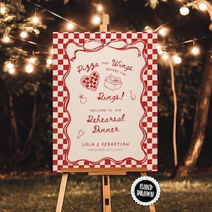 May include: A red and white checkered sign on a wooden easel, with the text "Pizza and Wings Before the Rings! Welcome to our Rehearsal Dinner" in a decorative font. The sign also includes the names Lola & Sebastian and the date November 10th, 20XX.