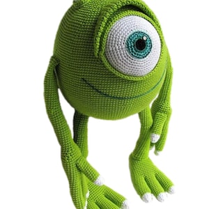 May include: A handmade, green crocheted plush toy of a one-eyed monster character. The toy has a large, round eye with a white iris and a blue-green pupil. It has small horns and arms and legs with white tips.