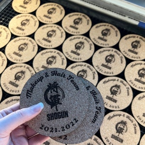 May include: Close-up of two round cork coasters with a black and white logo that says "SHOGUN" and "Makeup & Hair Team" with the year "2021.2022" printed on them.