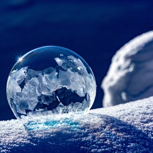 Może przedstawiać: Przezroczysta, zamrożona bańka z misternymi wzorami kryształków lodu spoczywa na śniegu. Bańka odbija światło, tworząc efekt iskrzenia na tle głębokiego błękitu. Obraz uchwyca piękno zimy.