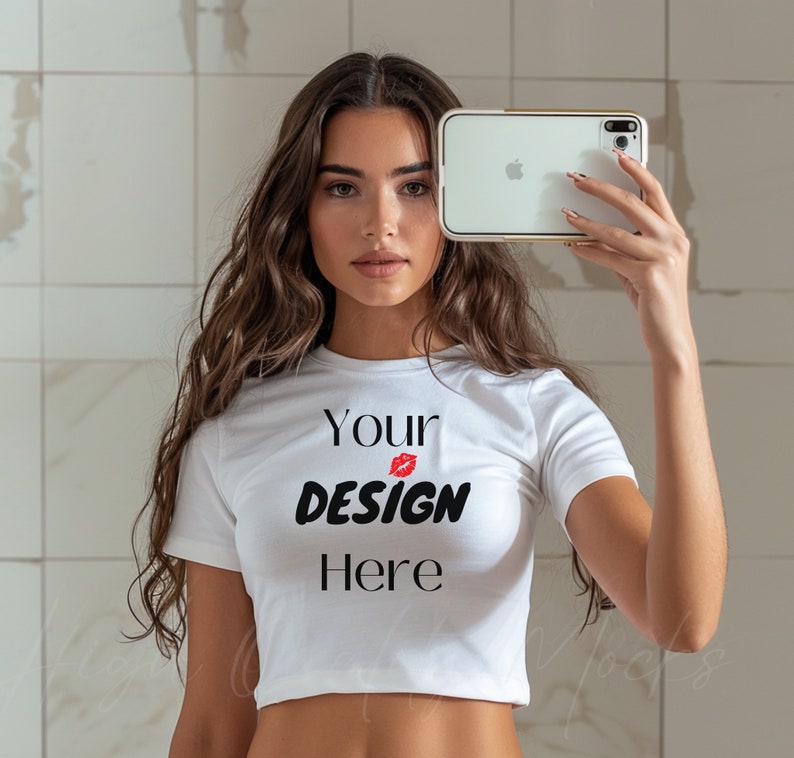 May include: A woman wearing a white crop top with the text "Your Design Here" and a red lip print. She is holding a white smartphone in front of her face.