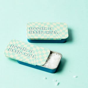 May include: Two small blue and white checkered tins with the text "needs a little salt" on the lid. One tin is open and contains white salt.